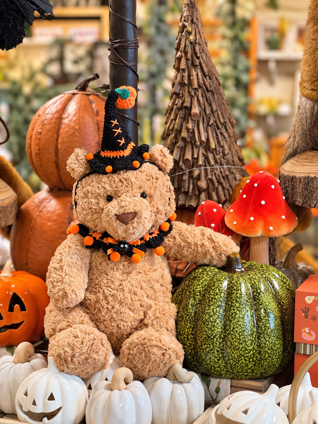 Brown plush bear wearing a black and orange Halloween witch hat and matching collar surrounded by pumpkins