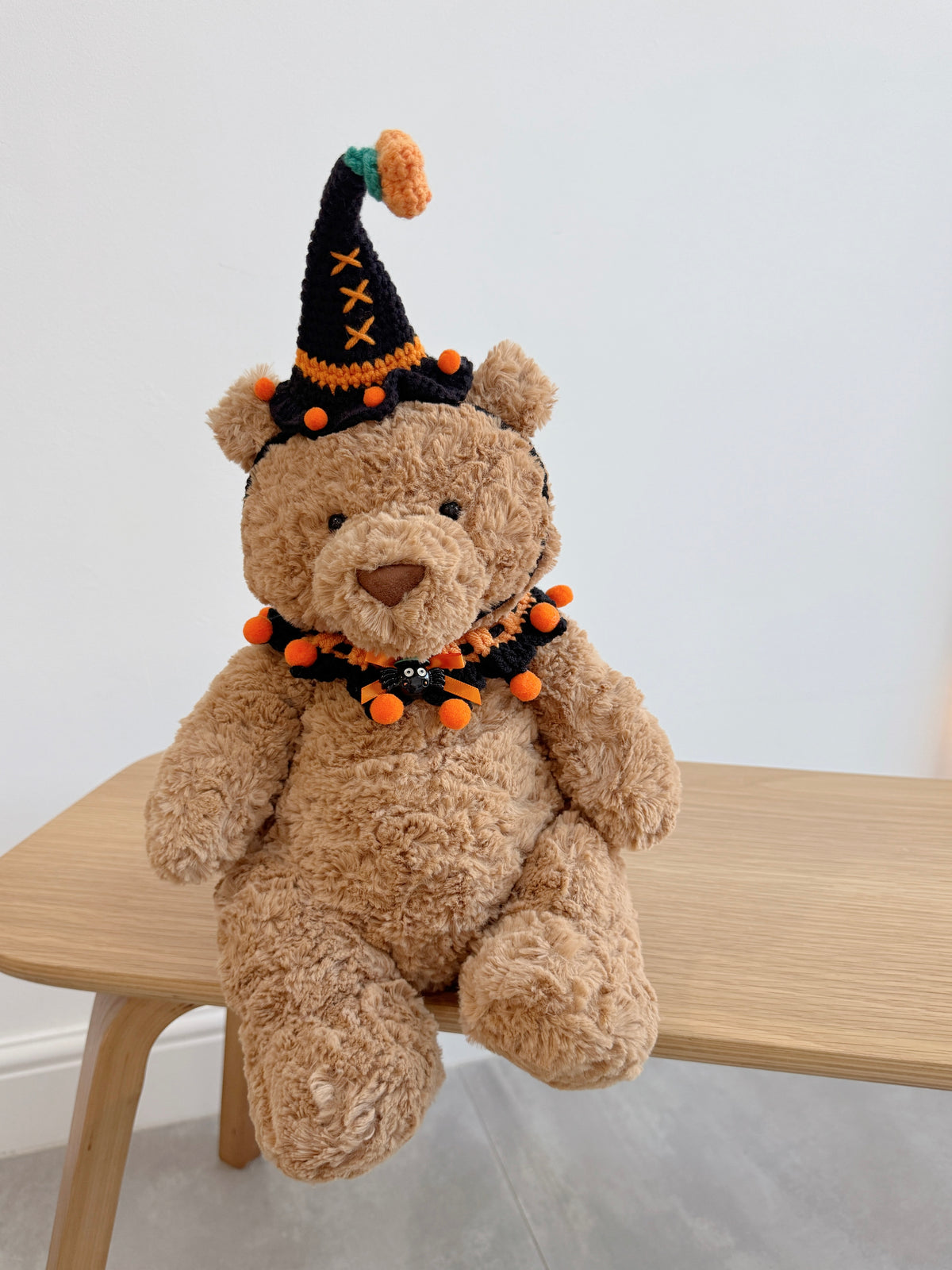 A plush brown teddy bear wearing a crochet black pumpkin hat and orange ruffled Halloween collar