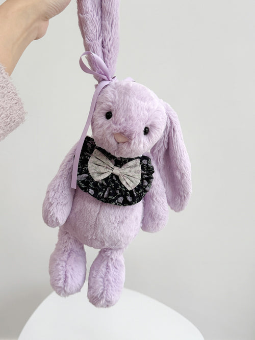 Purple plush bunny wearing a black floral ruffle bib with contrasting floral bow and adjustable lace ties